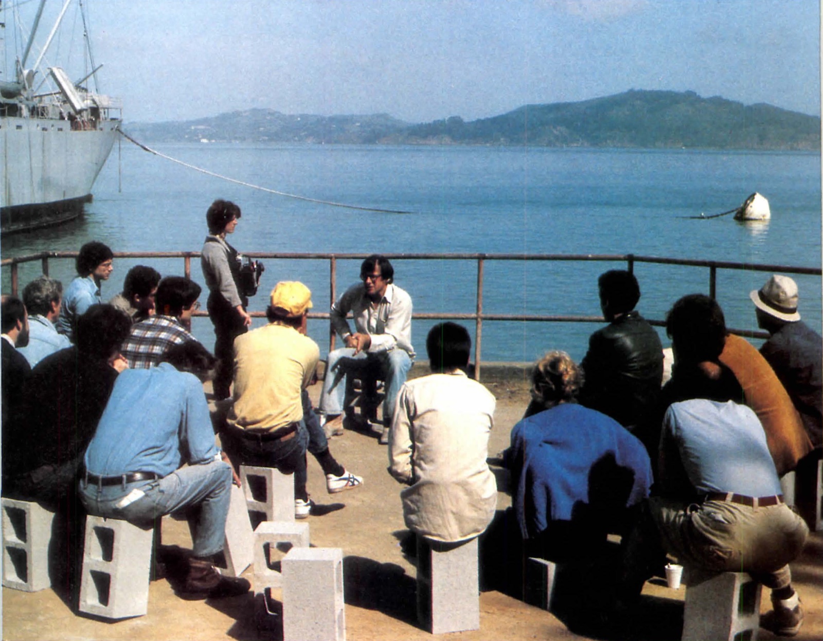 Christopher Alexander speaking to workshop participants seated on concrete blocks at the water’s edge at Fort Mason, with San Francisco Bay behind them, 1984.
